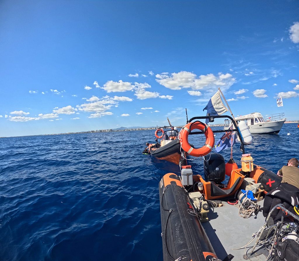 Two RAF Brize Norton Sub Aqua Club rigid inflatable rescue boats floating on calm blue ocean waters under a clear sky, equipped with orange lifebuoy rings, ropes, and flags, with another white boat is visible in the distance.
